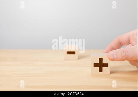 Blocco di segno di Person Holding Plus contro Minus. Più o meno. Uomo tenere cubo con più icona. Mano scegliere cubo di legno con segno più. Cubo di legno sfocato w Foto Stock