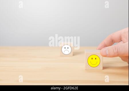 Mano scegliere blocco cubo di legno con icona faccia sorriso. L'uomo tiene il dito su un cubo di legno con faccia positiva. Cubo con segno di faccia negativa icona è Foto Stock