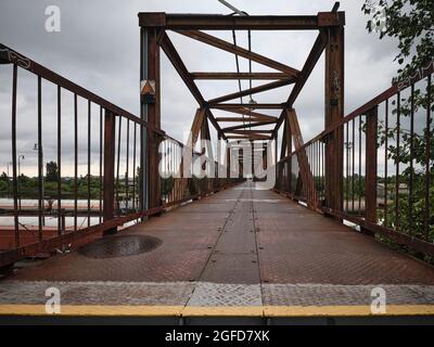 Ponte pedonale in ferro arrugginito con forti linee guida e prospettiva industriale sotto un cielo nuvoloso. Foto Stock