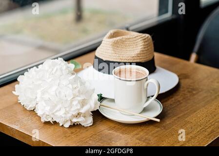 Una tazza di fragrante cioccolata calda con fiori bianchi di ortangea e un cappello sul tavolo in una caffetteria. Messa a fuoco selettiva soft. Foto Stock