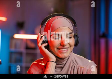 Primo piano ritratto di bella sorridente felice rilassata 30-età araba donna in hijab in cuffie ascoltare musica orientale tradizionale con occhi chiusi. Interni domestici di sera, luci notturne colorate Foto Stock