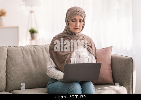 Grave femmina islamica di mezza età che usa il laptop che lavora in linea a casa Foto Stock