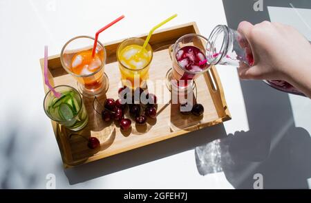 Un vassoio di legno con bevande colorate: Limonata arancione, gialla e cetriolo. Nelle vicinanze si trovano le ciliegie. Una mano versa il succo rosso da un decanter in un bicchiere. C Foto Stock