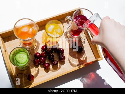 Un vassoio di legno con bevande colorate: Limonata arancione, gialla e cetriolo. Nelle vicinanze si trovano le ciliegie. Una mano versa il succo rosso da un decanter in un bicchiere. C Foto Stock