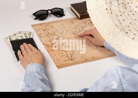 parte di una ragazza in un cappello di paglia. la mano tiene un portafoglio nero di pelle con soldi su un bianco occhiali da sole da tavola di legno, mappa del mondo e diario. fuoco selettivo Foto Stock