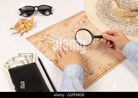 le mani femminili tengono una lente d'ingrandimento sulla mappa del mondo. su un tavolo di legno bianco giace un portafoglio nero con soldi, un cappello di paglia, una conchiglia e occhiali da sole. Foto Stock