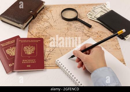 la mano femminile fa note con una matita in un taccuino. su un tavolo di legno bianco si trova una mappa del mondo, due passaporti, una lente d'ingrandimento e una wa nera Foto Stock