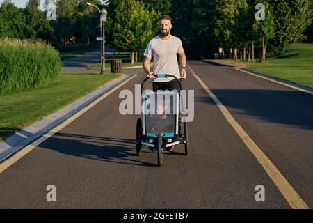 Giovane padre con il bambino in un passeggino da jogging durante il jogging in un parco pubblico. Togetherness papà con figlio durante la corsa attiva, paternità felice Foto Stock