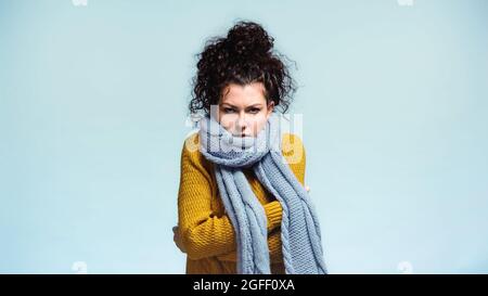 donna in caldo maglione e sciarpa che si abbraccia mentre gelida isolato su blu Foto Stock