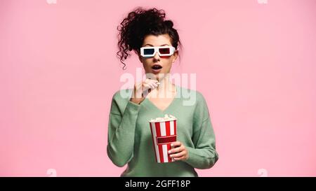 donna stupita in 3d occhiali che tengono secchio di popcorn isolato su rosa Foto Stock