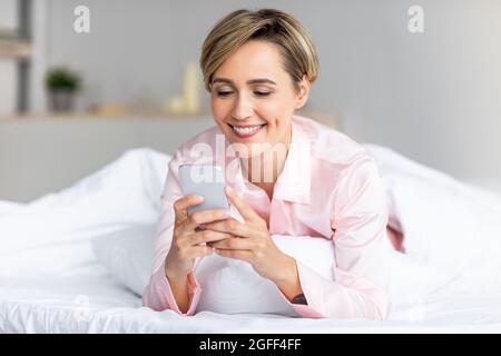 Donna felice sdraiata a letto leggendo il messaggio di testo al telefono Foto Stock
