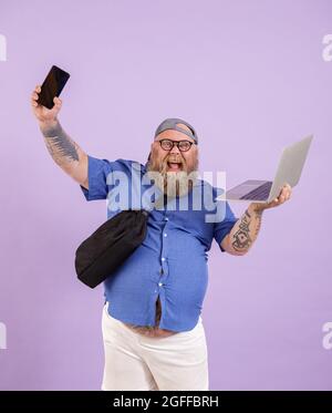 Un uomo positivo e di dimensioni più grandi con laptop e borsa mostra il telefono su sfondo viola in studio Foto Stock