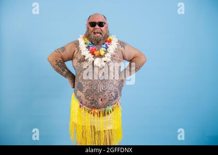 Uomo felice con sovrappeso e pancino tatuato in gonna decorativa in erba su sfondo azzurro chiaro Foto Stock