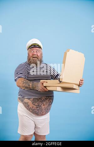 Felice sorpreso uomo marinaio plump tiene aperta scatola di pizza su sfondo azzurro chiaro Foto Stock