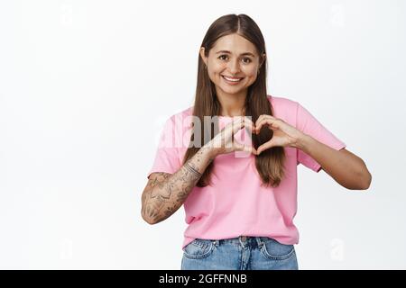 La ragazza romantica mostra il segno del cuore e sorride alla macchina fotografica, si erge in una maglietta rosa con jeans, sfondo bianco Foto Stock