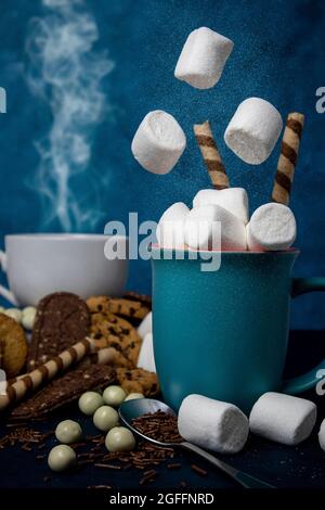 Marshmallow cade in una tazza circondata da vari biscotti e una tazza fumante Foto Stock