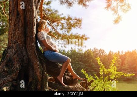 Giovane donna incinta siede sulle radici sotto un albero vicino al fiume Foto Stock