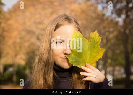 Bella giovane donna su uno sfondo di alberi autunnali. Concetto di emozione. La ragazza tiene in mano una foglia di acero e copre un occhio con la foglia. Foto Stock