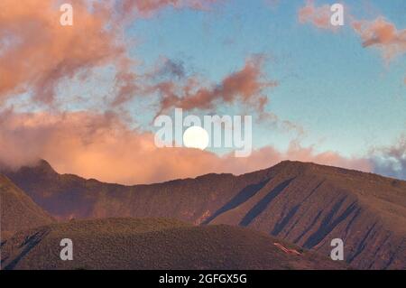 Luna piena che si innalza sulle montagne ovest di maui al tramonto con nuvole rosa puffy. Foto Stock