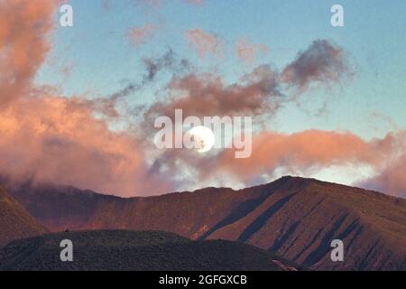 Luna piena che si innalza sulle montagne ovest di maui al tramonto con nuvole rosa puffy. Foto Stock