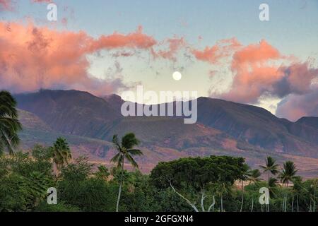 Luna piena che si innalza sulle montagne ovest di maui al tramonto con nuvole rosa puffy. Foto Stock