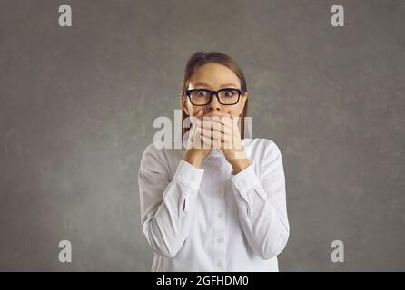 Giovane donna scioccata in occhiali con mano sopra bocca studio ritratto scatto Foto Stock