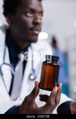 Primo piano di mano afroamericana con bottiglia di pillole per il trattamento raccomandato. Matraccio di contenimento medico nero con medicinali, medicamenti e farmaci. Medico con fiala in armadietto medico Foto Stock