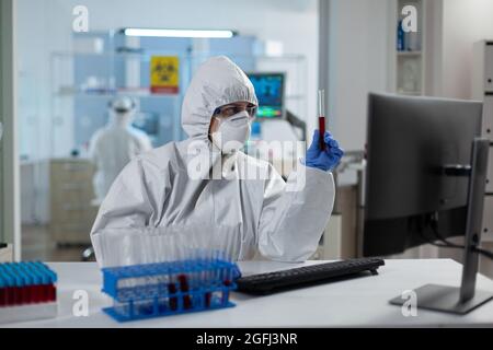 Donna ricercatrice biologa che tiene in mano la provetta per l'analisi delle competenze in materia di liquidi lavorando allo sviluppo del vaccino del coronavirus in laboratorio. Scienziato che indossa il vestito ppe durante l'esperimento medico Foto Stock