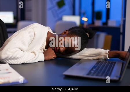 Stanco esausta donna d'affari afroamericana che dorme sul tavolo della scrivania a tarda notte in ufficio di startup dopo aver lavorato alle statistiche di gestione. Workaholic black manager pianificazione strategia aziendale Foto Stock