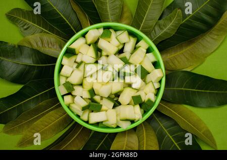 Pezzi di mango crudi in una ciotola verde con foglie di mango su sfondo verde Foto Stock
