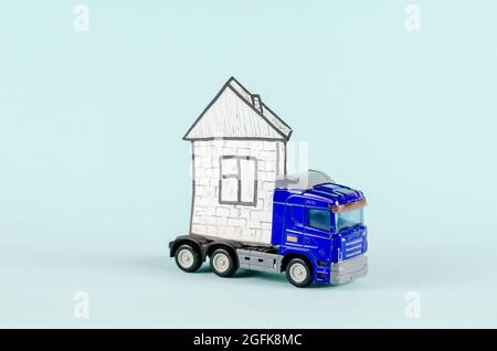 Trattore-rimorchio giocattolo che trasporta una casa. Disegno di casa e camion in plastica blu su carta bianca. Sfondo blu. Immobiliare, industria, trasporti. SEL Foto Stock