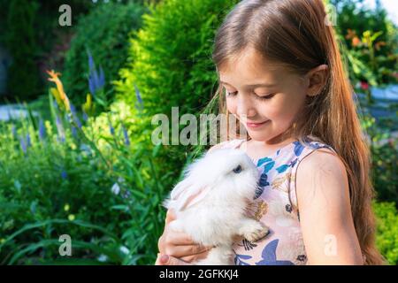 Una ragazza carina sta tenendo un coniglietto bianco del bambino sul suo petto. Un coniglio nana decorativo nelle mani di una ragazza bionda. Il concetto di amicizia tra un c Foto Stock