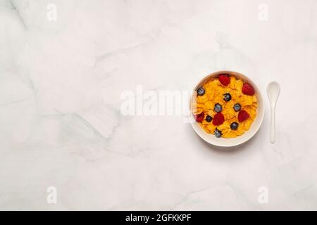 La colazione del mattino comprende una ciotola di cereali con fiocchi di mais su una superficie di marmo bianco con lamponi e mirtilli in una ciotola bianca con cucchiaio bianco. Spazio di copia Foto Stock