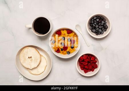 La colazione del mattino comprende cereali al fiocco di mais con lamponi freschi e mirtilli con bagel e formaggio spalmabile con caffè nero Foto Stock