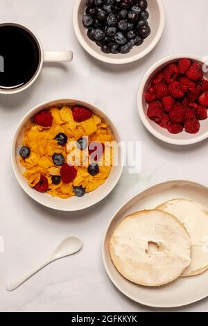 La colazione del mattino comprende cereali al fiocco di mais con lamponi freschi e mirtilli con bagel e formaggio spalmabile con caffè nero Foto Stock
