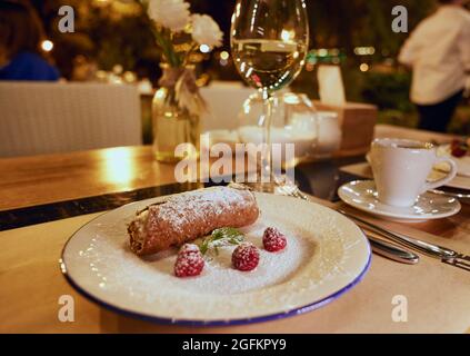 I cannoli siciliani rotolano con mirtilli su un piatto di ceramica bianca e un bicchiere di vino bianco su un tavolo da ristorante con un giardino cittadino sullo sfondo. Foto Stock