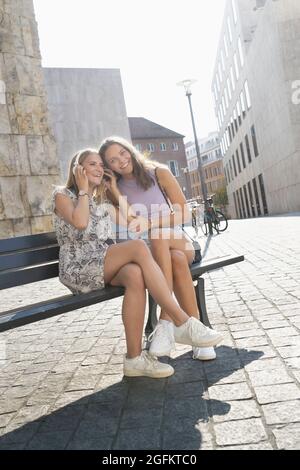 Due giovani donne che ascoltano musica con cuffie su una panchina in città Foto Stock