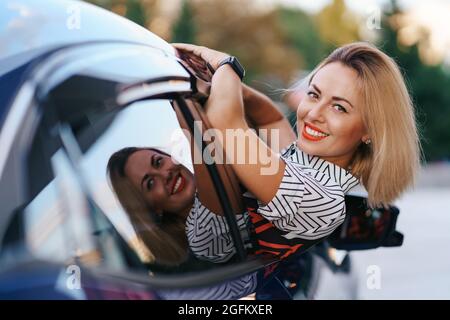 Allegra donna caucasica guida attraverso la pittoresca città soleggiata e. ondeggia le braccia mentre si allunga fuori dal finestrino dell'auto in una bella giornata Foto Stock