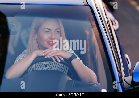 Ritratto di signora in auto. Giovane bella donna che guida. Vista frontale attraverso il parabrezza con luce solare. Foto Stock