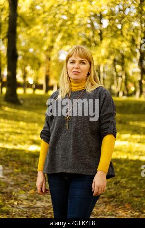 Defocalizzare la donna bionda seria anni '40 in piedi nel parco autunno giallo. Felice bella signora. Donna con pullover grigio, dolcevita giallo, pendente, jeans. Foto Stock