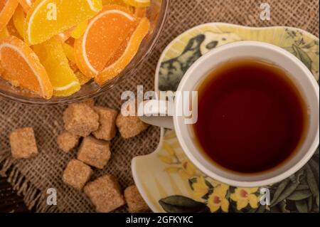 Fette di limone di marmellata multicolore in vaso di vetro, pezzi di zucchero di canna di canna e una tazza di tè su un piatto, primo piano, fuoco selettivo Foto Stock