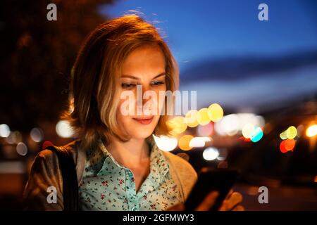 Primo piano ritratto di una giovane donna utilizzando uno smartphone su uno sfondo di notte luci della città Foto Stock