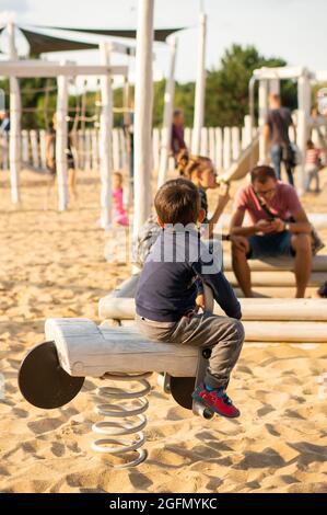 POZNAN, POLONIA - 26 ago 2018: Un bambino che indossa un maglione blu seduto su attrezzature di legno swing sulla sabbia in un parco giochi Foto Stock