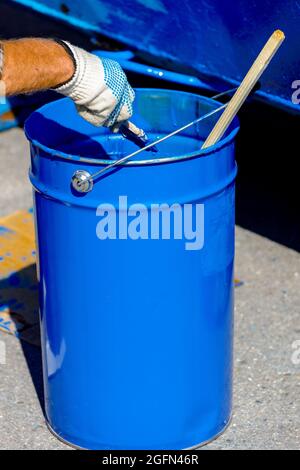 Secchio in metallo con vernice blu. Una mano in un guanto di protezione tiene una spazzola di vernice. Verniciatura di prodotti metallici. Foto Stock
