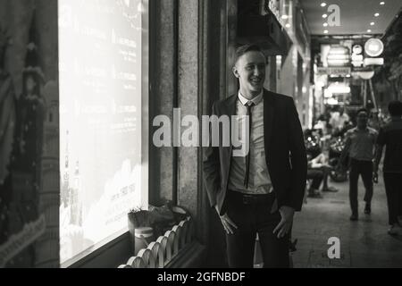 Ritratto in bianco e nero di giovane uomo d'affari attraente in elegante abito formale a piedi sulla strada con le luci nella grande città. Foto notturna. Foto Stock