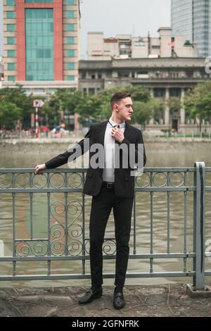 Bel giovane uomo in abito nero formale in piedi vicino al fiume nella grande città. Raddrizza la cravatta. Grattacieli sullo sfondo Foto Stock