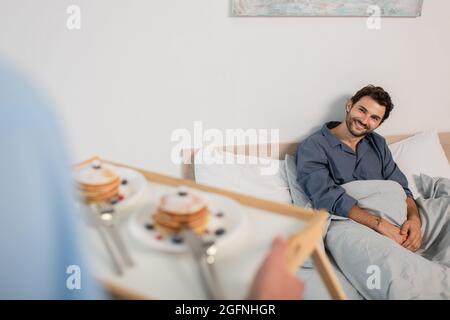 uomo sfocato che tiene vassoio per la colazione con frittelle vicino ragazzo felice a letto Foto Stock