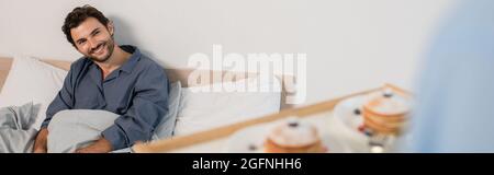 uomo sfocato che tiene vassoio per la colazione con frittelle vicino ragazzo felice a letto, banner Foto Stock