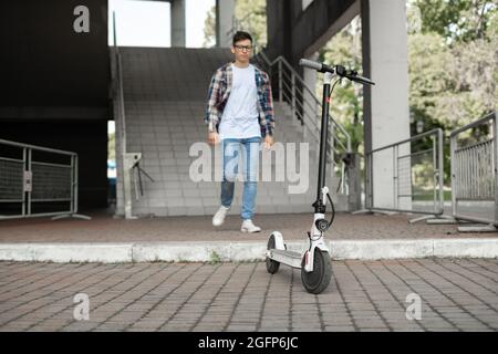 Un uomo attraente esce dal centro commerciale e si dirige verso uno scooter elettrico parcheggiato. Vista dal retro senza volto. Stile di vita tecnologico ecologico. Foto Stock