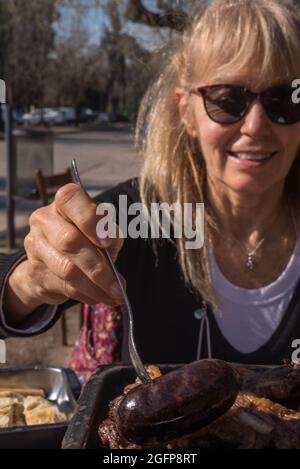 Donna adulta che pica una salsiccia di sangue con la sua forchetta da un barbecue argentino. Messa a fuoco in primo piano Foto Stock